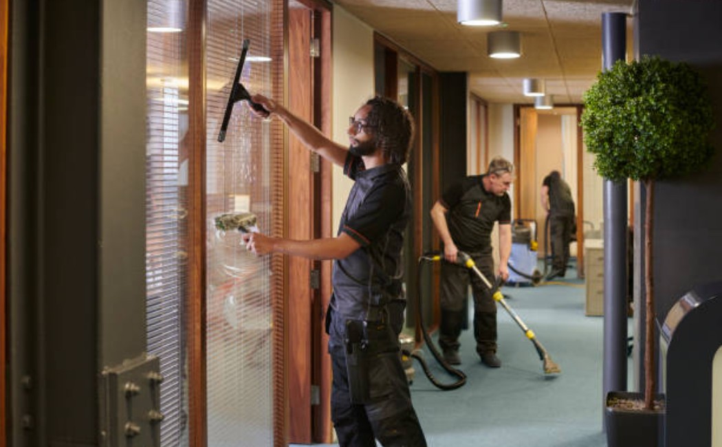Professional cleaners in safety vests are cleaning an office workspace.